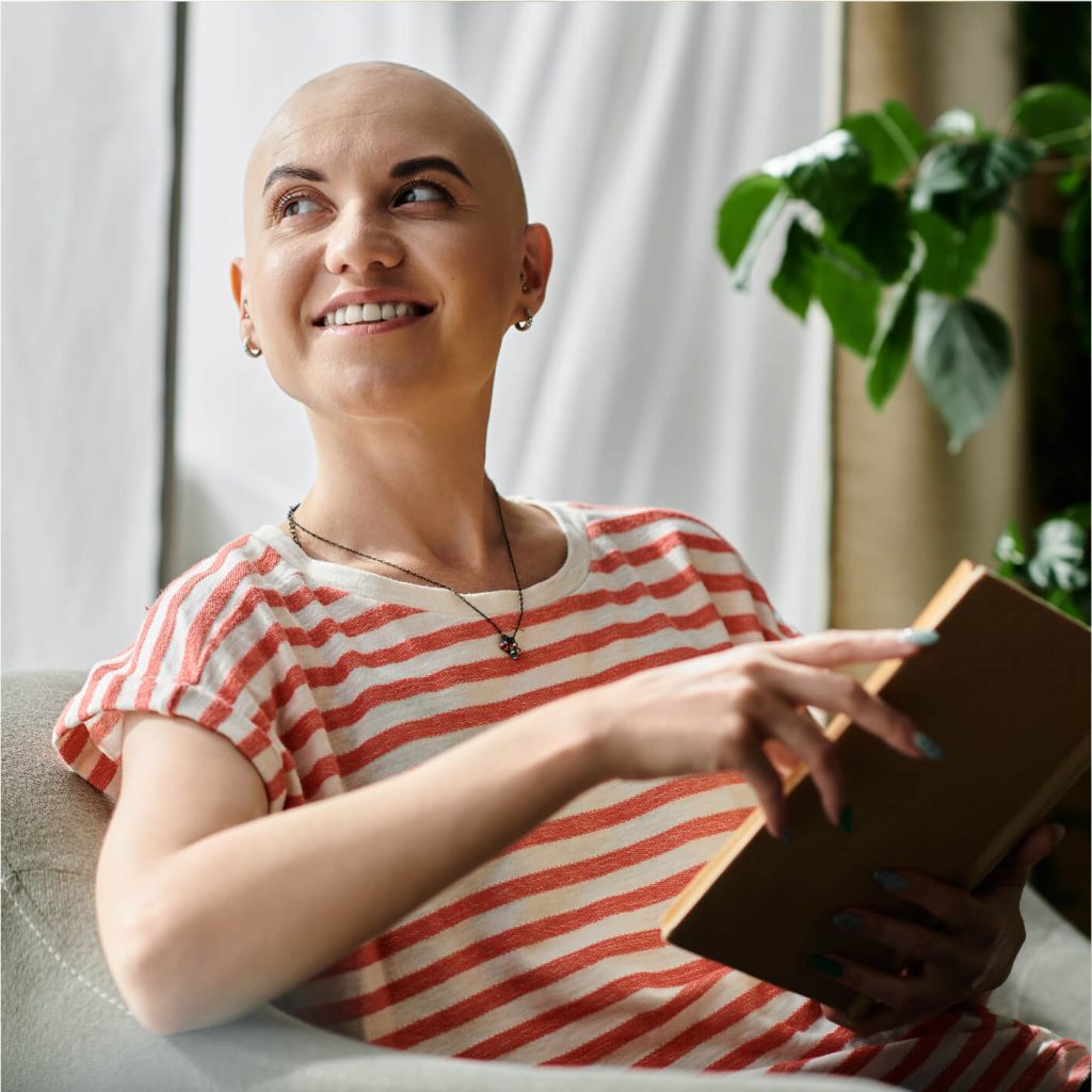 A smiling person with a bald head, wearing a striped shirt, sits indoors.