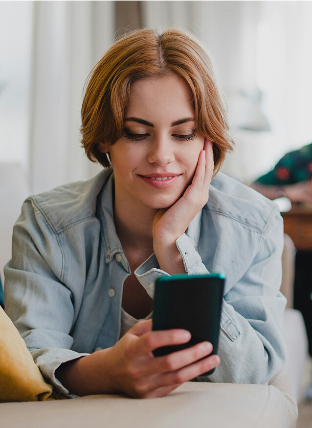 A woman with short light brown hair in a denim shirt is lying on a couch, smiling while looking at her smartphone.