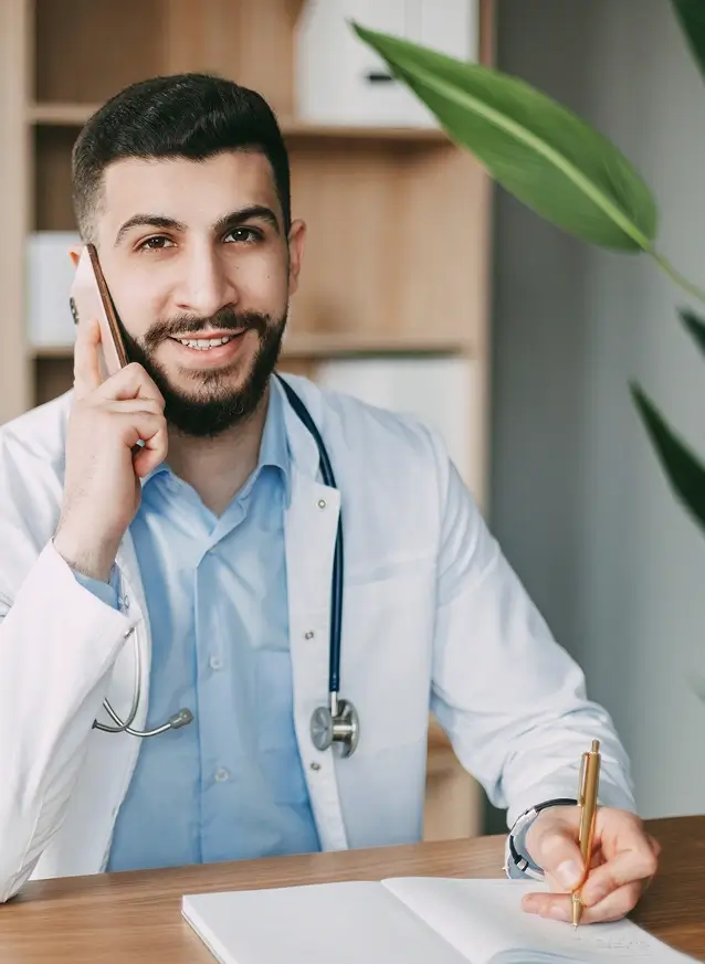 A male doctor in a white coat with a stethoscope talks on the phone.